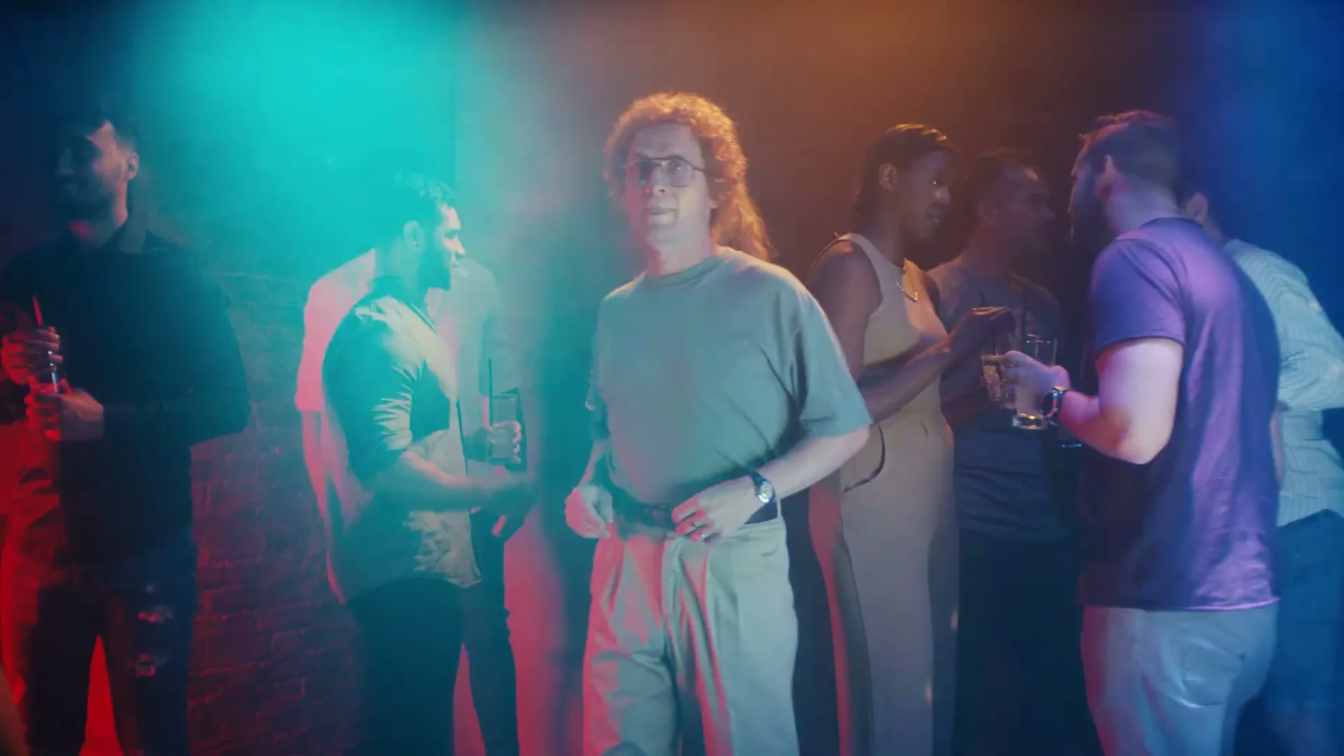 A man with curly hair and glasses dances awkwardly in a dimly lit club, surrounded by other partygoers holding drinks.