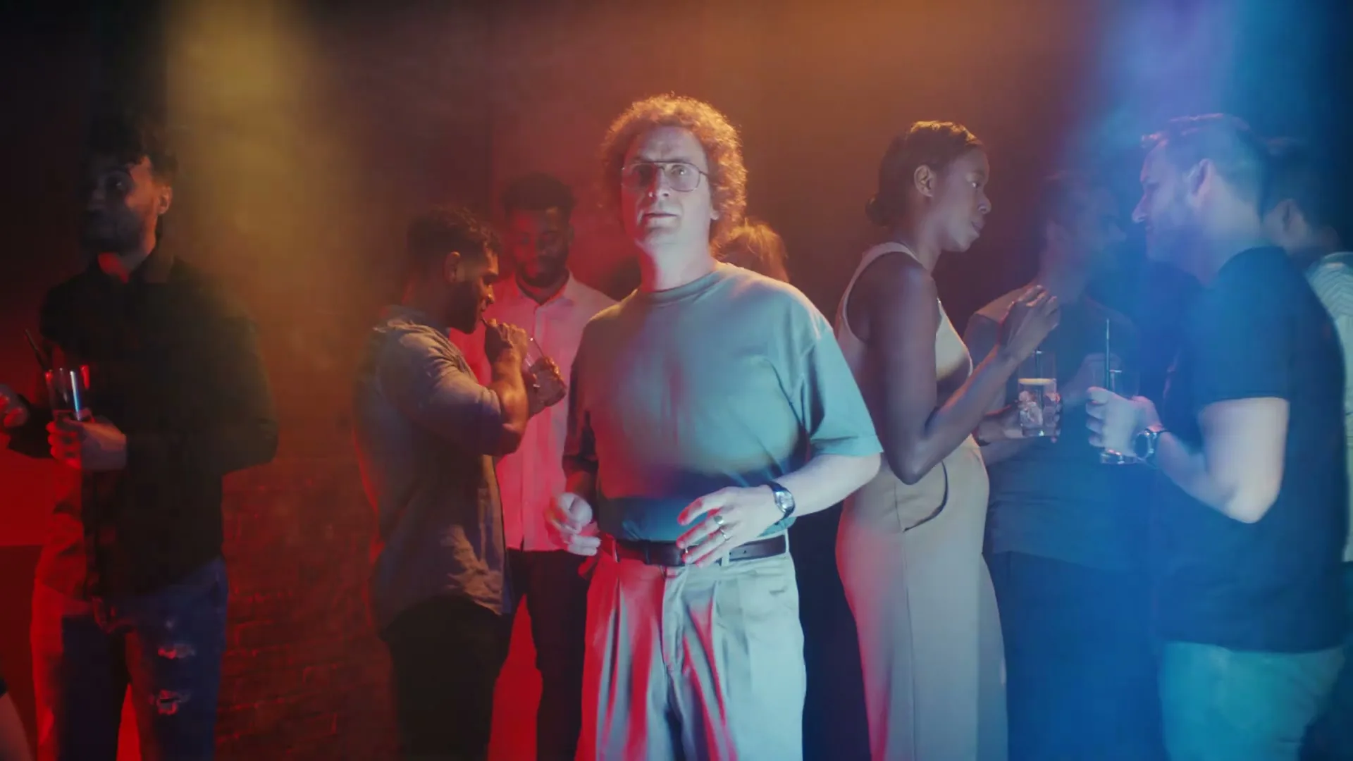 A man with curly hair and glasses stands awkwardly in a dimly lit, crowded club, surrounded by other partygoers.