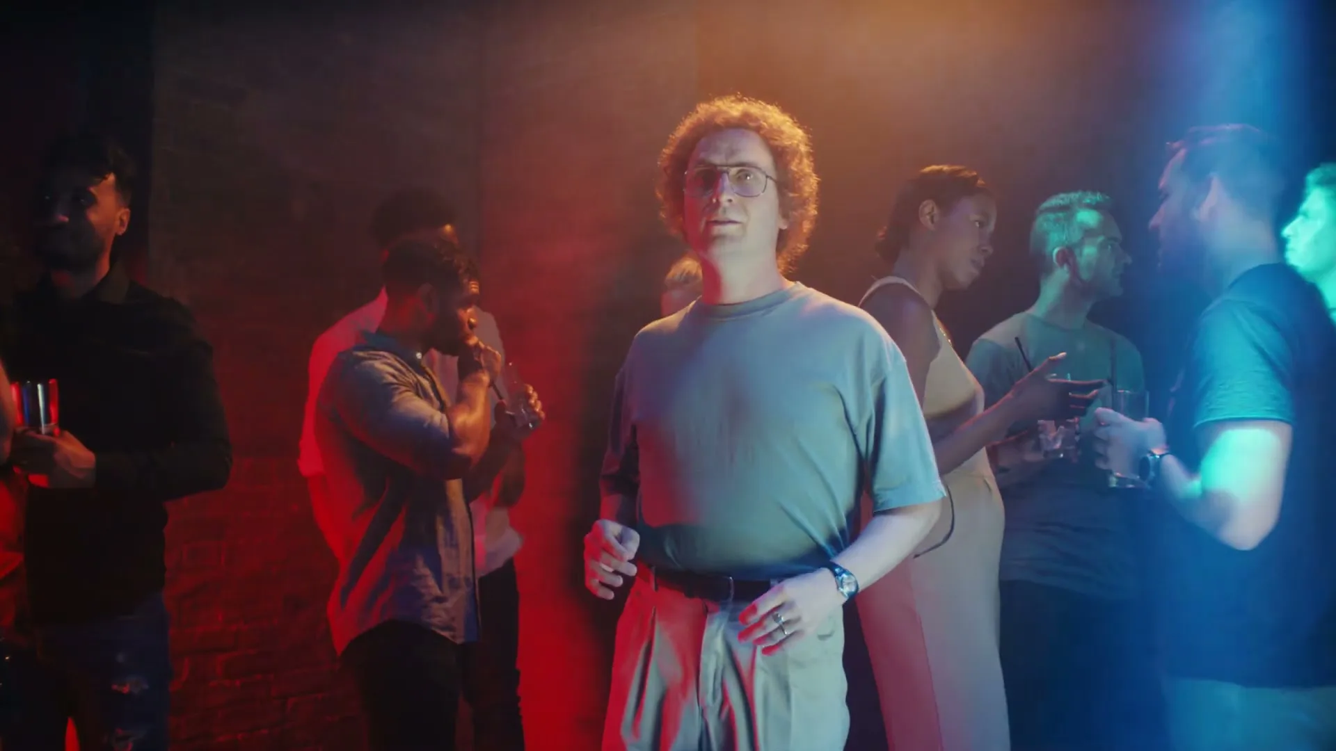 A man with curly hair and glasses stands in a crowded, dimly lit club, looking around with a slightly confused expression.