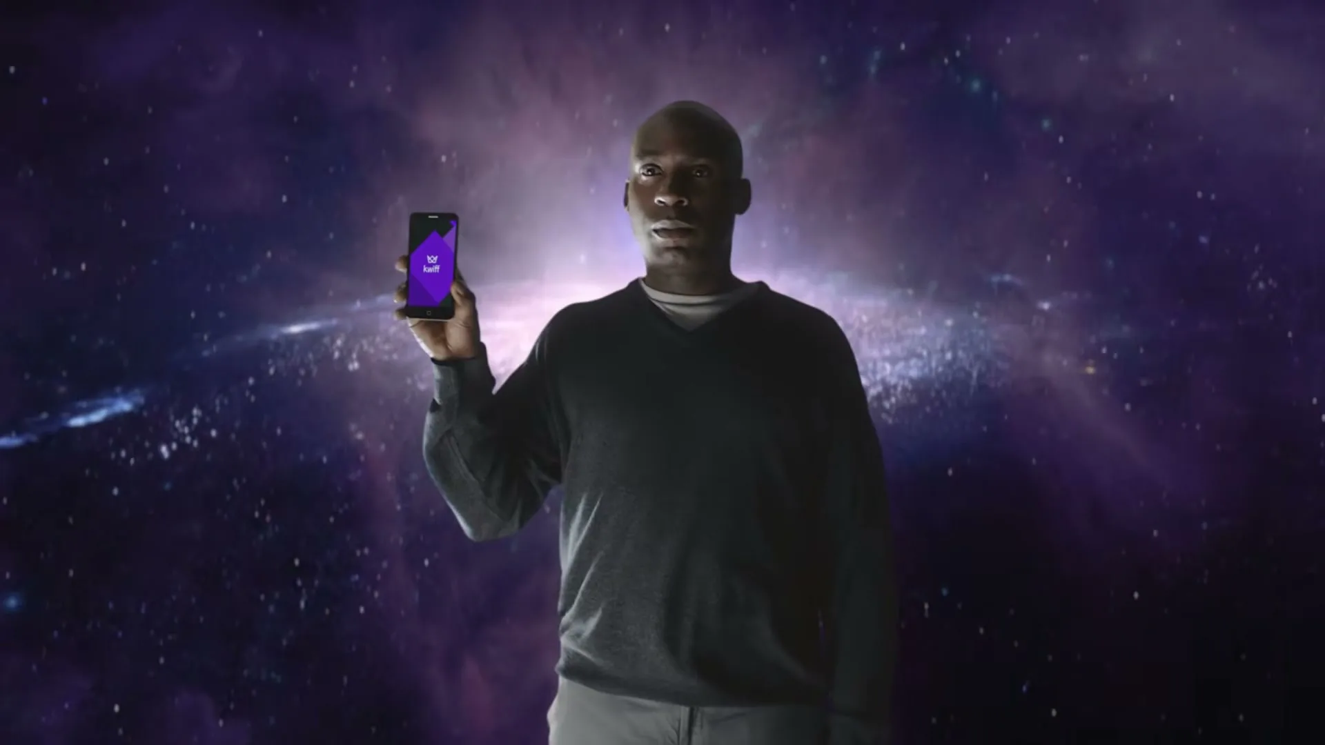A Black man in a dark sweater holds up a smartphone with a purple screen against a backdrop of a purple galaxy.