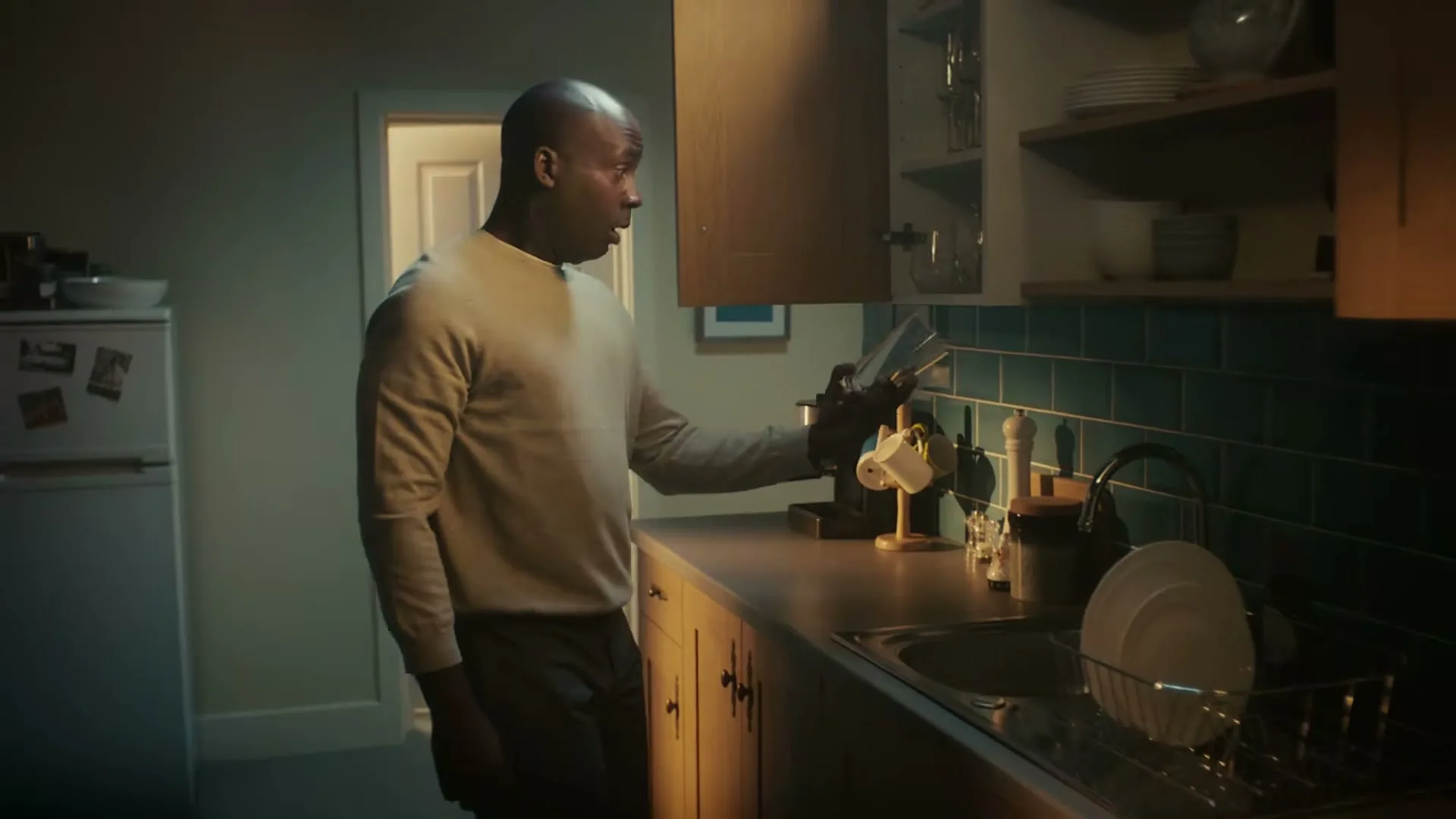 A man in a beige sweater stands in a kitchen, opens a cabinet, and catches a falling glass before it hits the ground, looking surprised.