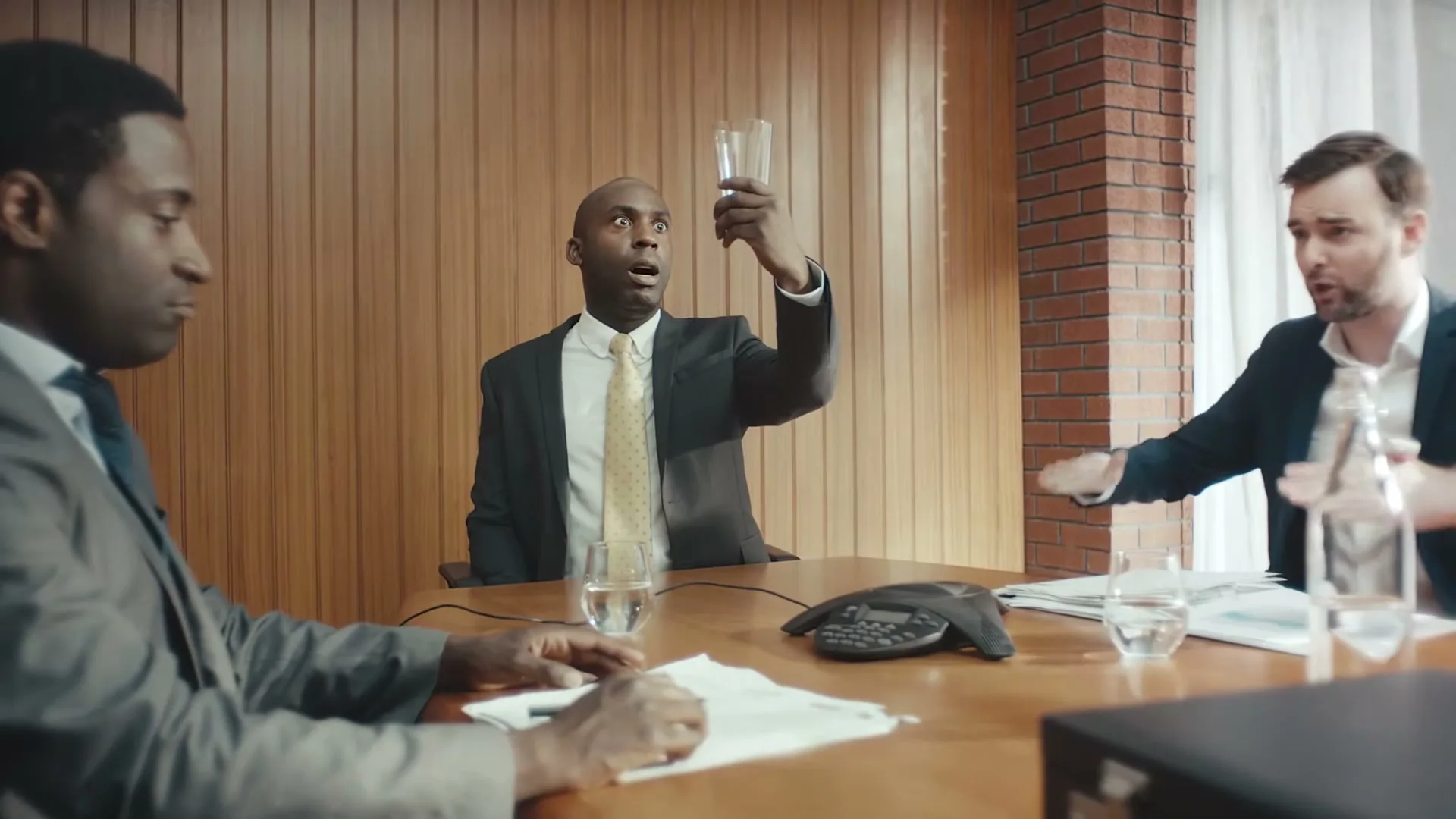 Three businessmen in suits sit around a conference table, one holding a glass in amazement, while another gestures excitedly.