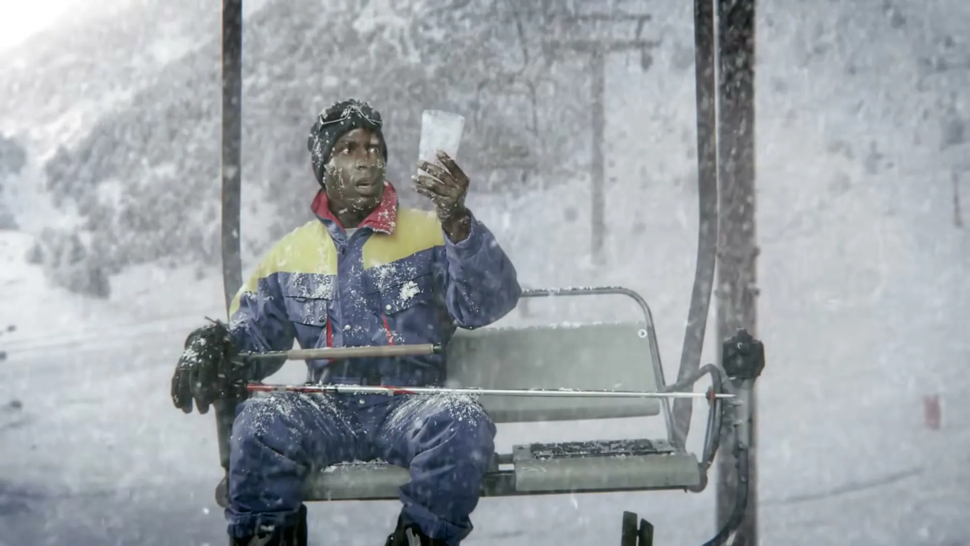 A man in a blue and yellow ski suit sits on a ski lift, covered in snow, holding a glass of ice in a snowy mountain.