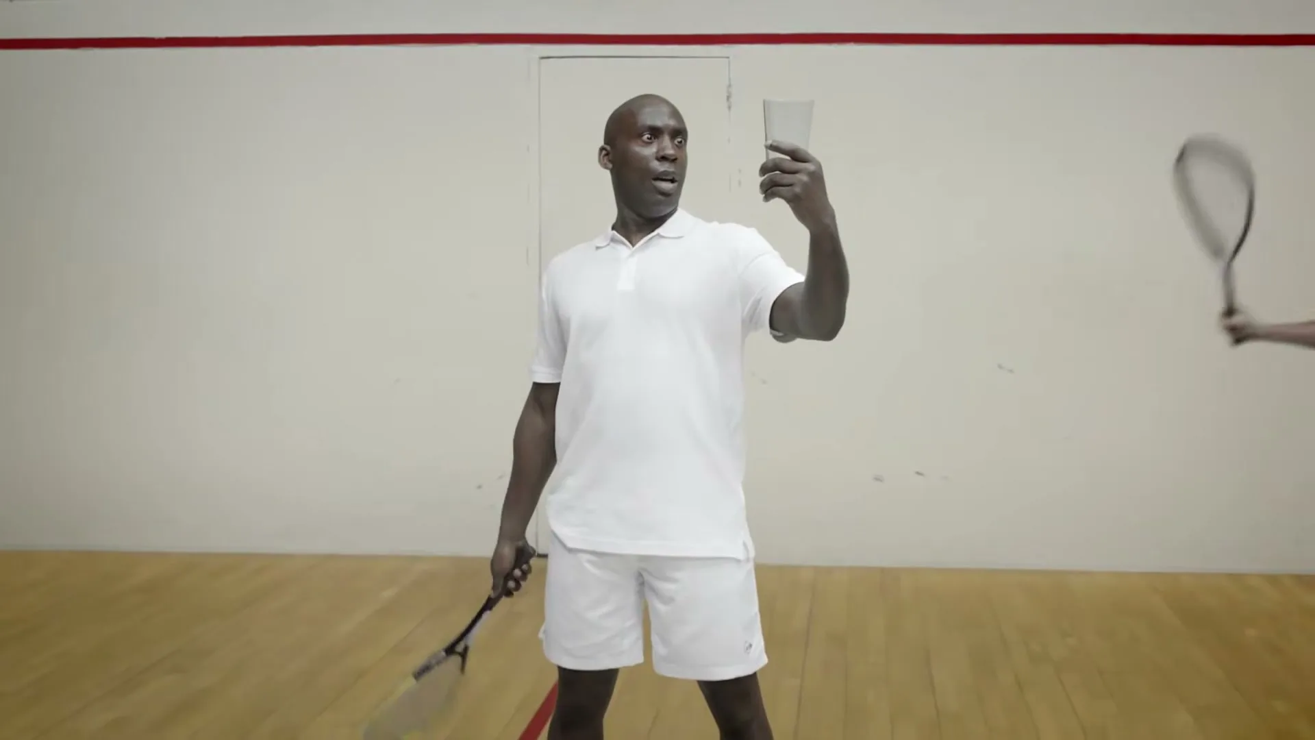 A man in white tennis attire holds a glass in the air, looking surprised, while another man swings a racket in a squash court.