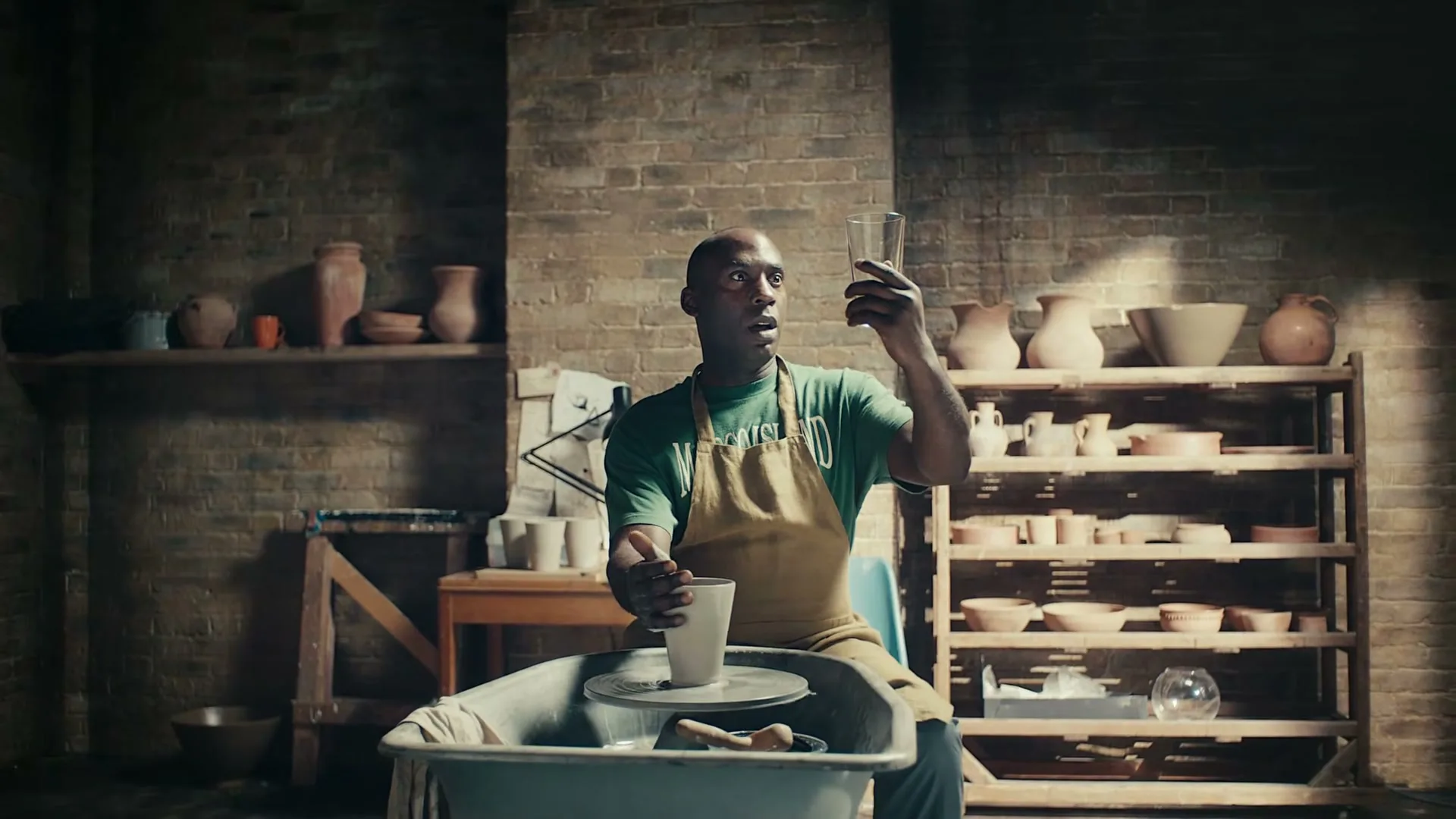 A man in a green shirt and apron sits at a pottery wheel, holding a glass up to the light and looking surprised in a rustic studio.