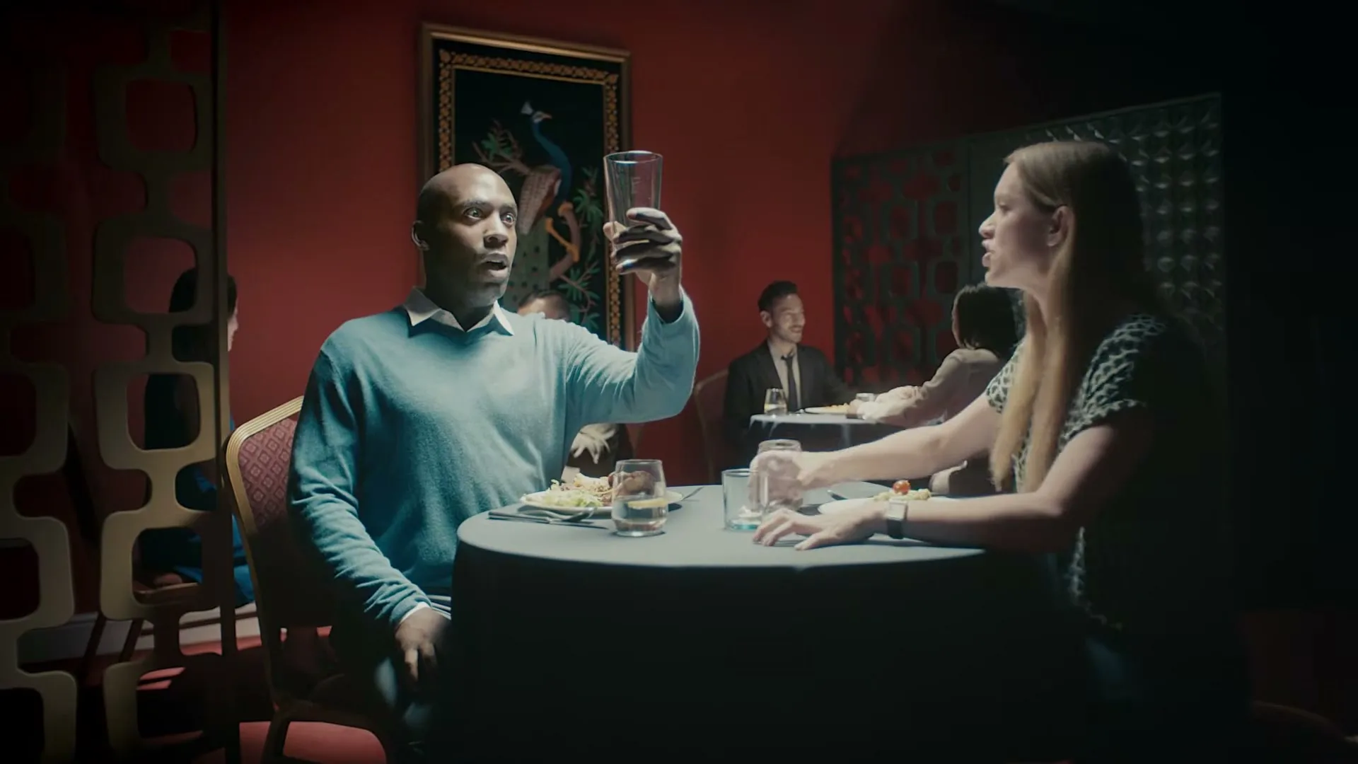 A man in a blue sweater holds up an empty glass, looking surprised, as a woman in a leopard print top stands up from their table in a dimly lit restaurant.