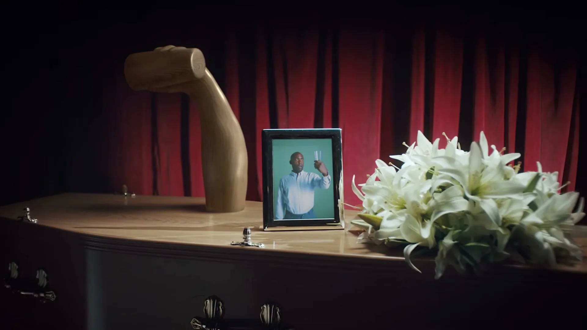 A framed photo of a man holding a glass is on a coffin, next to a bouquet of white flowers, in a dimly lit room with red curtains.