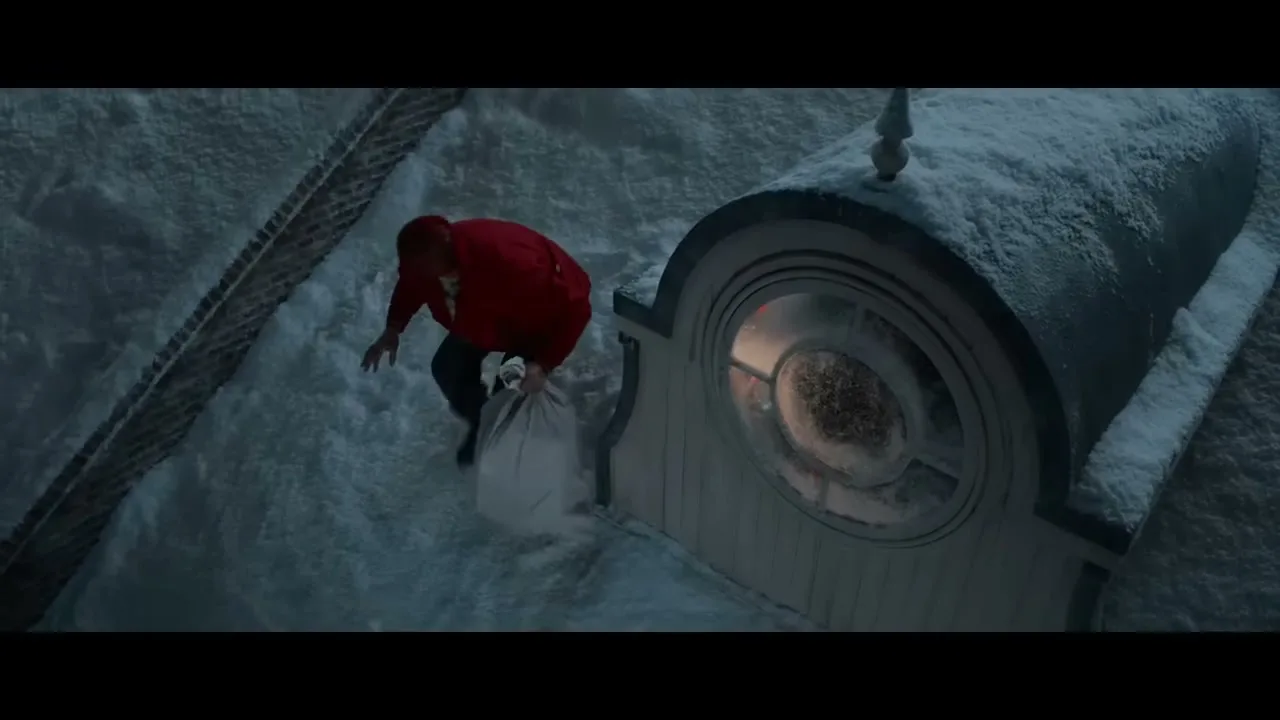 A person dressed in a red jacket and dark pants, resembling Santa Claus, struggles to climb onto a snow-covered roof next to a round window, holding a white sack.