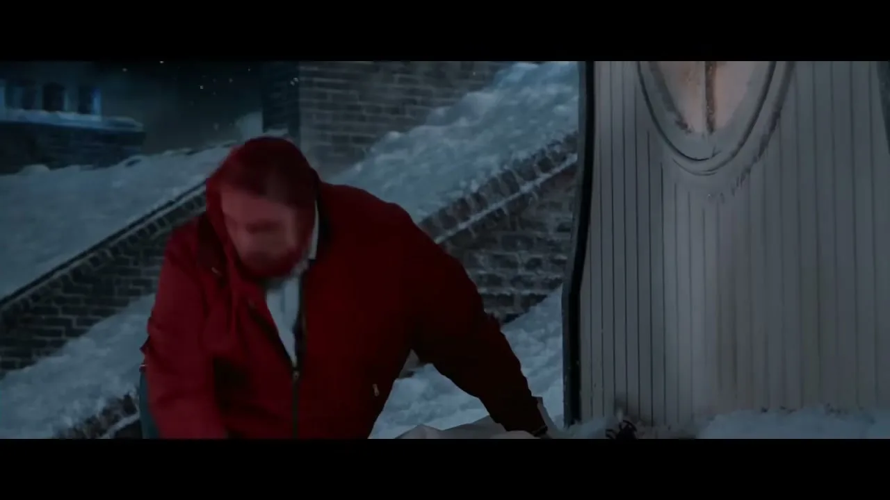 A man in a red jacket and blue jeans is on a snowy rooftop at night, looking down and searching for something.