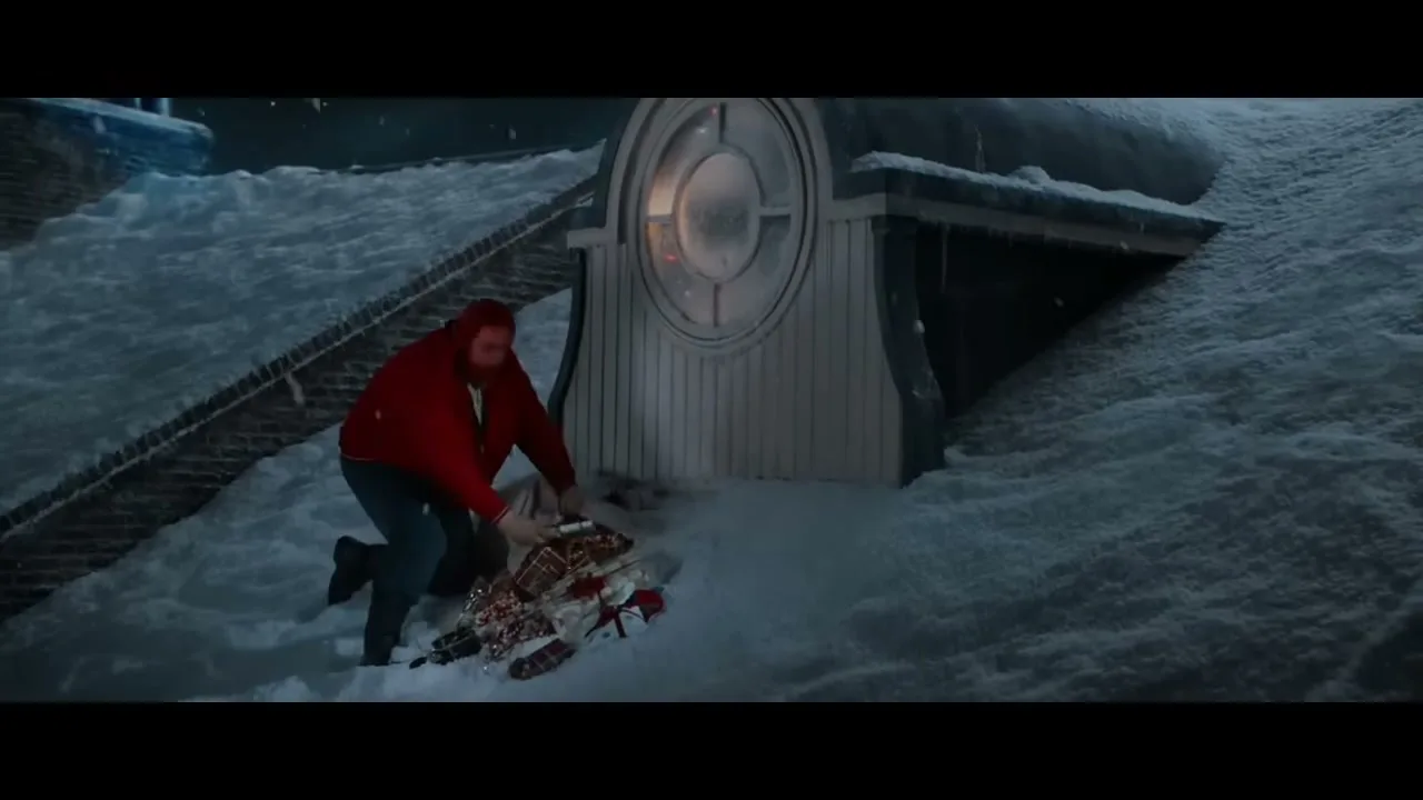 A man in a red jacket struggles to pull a sack of presents through deep snow on a rooftop at night.