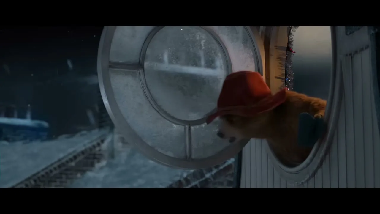 Paddington Bear, wearing a red hat and blue mittens, peeks out of a round, frosted window at a snowy rooftop at night.