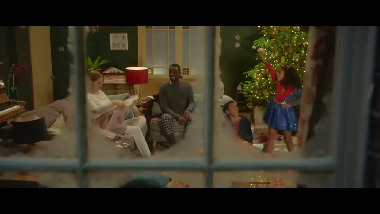 A family of four, including a young girl in a superhero costume, celebrates Christmas in their living room, seen through a frosted window.