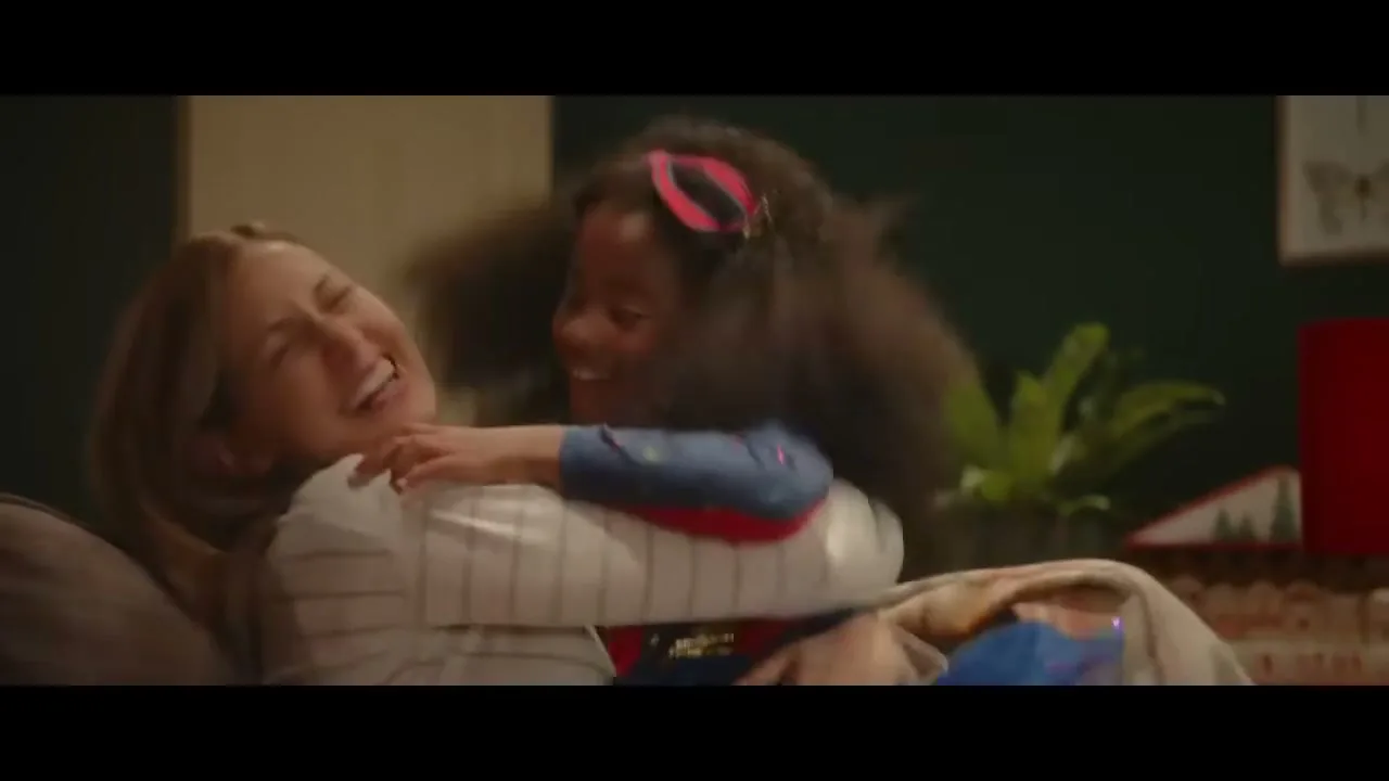 A young girl in a superhero costume jumps into the arms of a laughing woman, embracing her warmly in a festive living room with a Christmas tree.