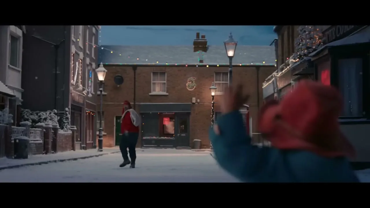 A man in a red hat and jacket waves goodbye to Paddington Bear, who is out of focus in the foreground, on a snowy street at dusk.
