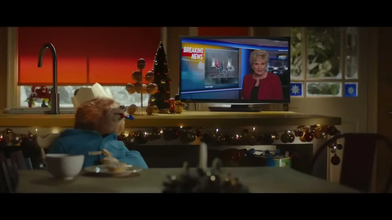 Paddington Bear, wearing a paper crown and blue coat, sits at a kitchen table watching a female news anchor on TV report on Santa.
