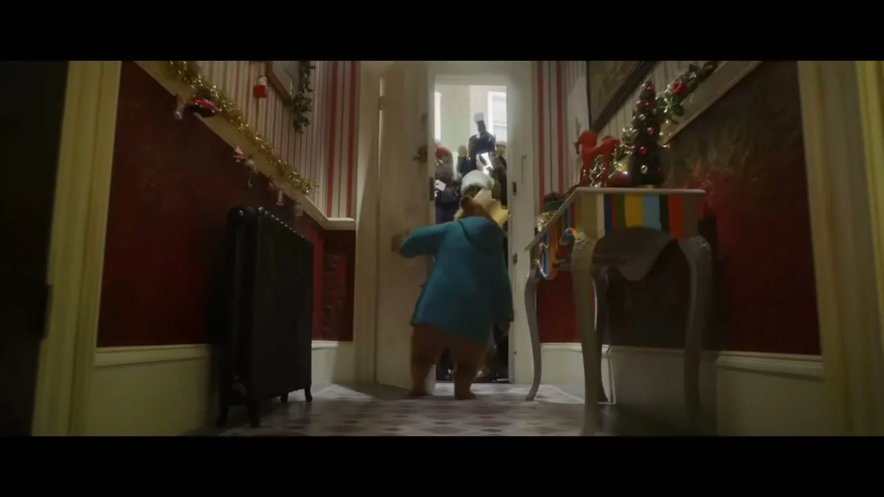 Paddington Bear, wearing a blue coat and red hat, opens the door to a group of carolers in a festive hallway.