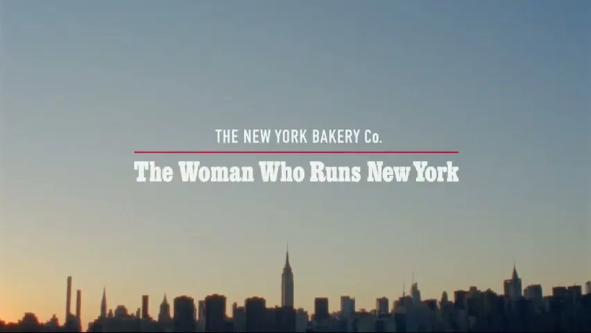 A wide shot of the New York City skyline at sunrise with the title card "The New York Bakery Co. The Woman Who Runs New York" overlaid.