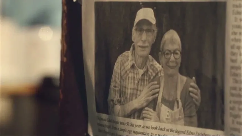A black and white newspaper photo of an elderly couple, a man in a cap and a woman in glasses, smiling and embracing, is shown on a wall.