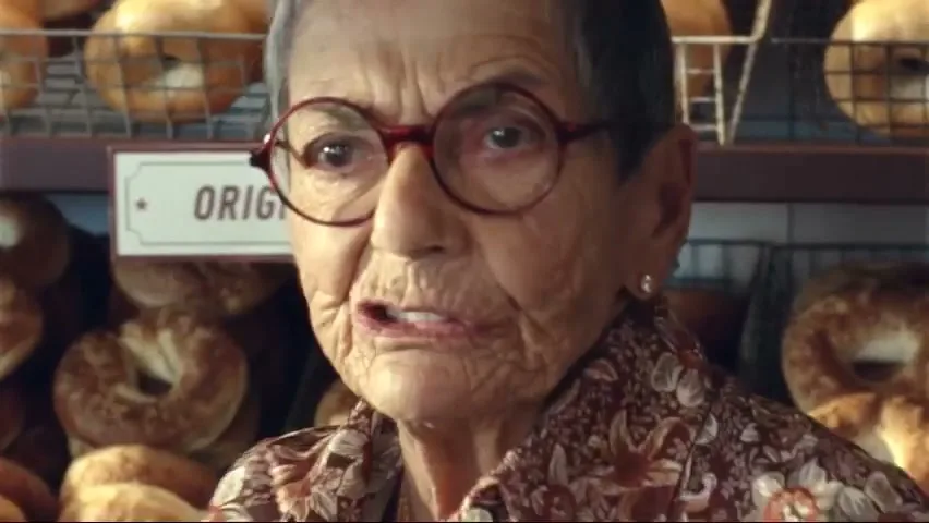 An elderly woman with short grey hair and red glasses turns to face the camera in a bagel shop, with shelves of bagels in the background.
