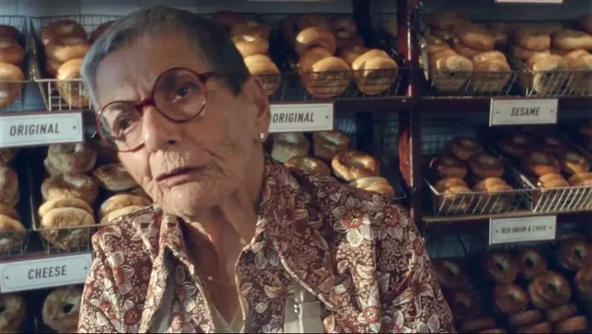 An elderly woman with short grey hair and glasses, wearing a patterned shirt, looks at the camera in a bagel shop.