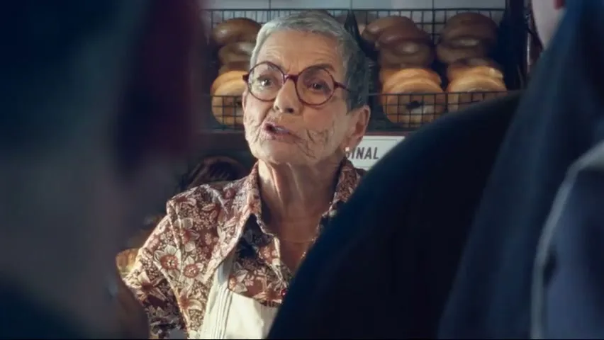 An older woman with short grey hair and glasses, wearing a patterned shirt and apron, speaks to someone off-camera in a bagel shop.