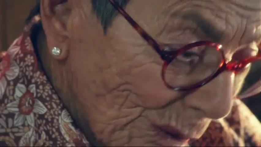 A close-up of an older woman with red glasses and a floral shirt, looking down with a slight smile, in an interior setting.