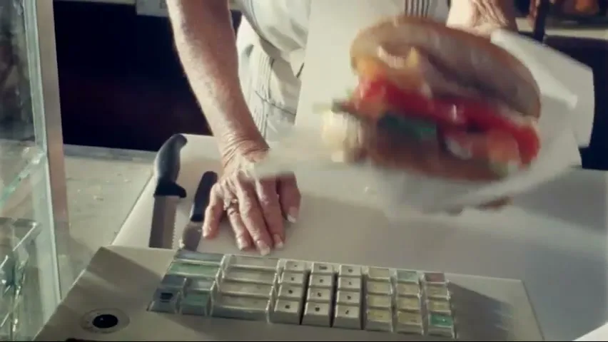 A woman's hands are seen placing a large sandwich wrapped in paper onto a counter next to a cash register.