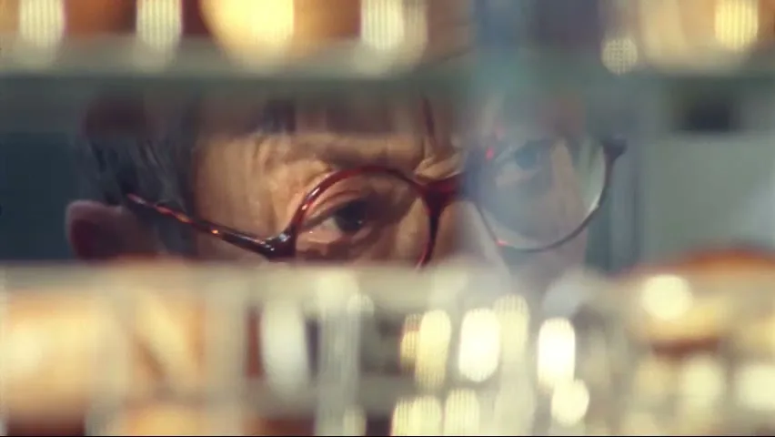 A woman with red glasses looks up from behind a blurry display of baked goods in a bakery.