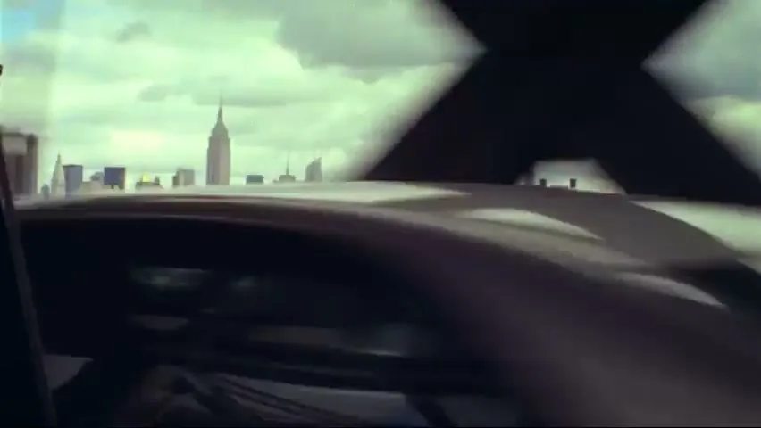 A wide shot of the New York City skyline with the Empire State Building visible, seen from a moving vehicle on a bridge on a cloudy day.