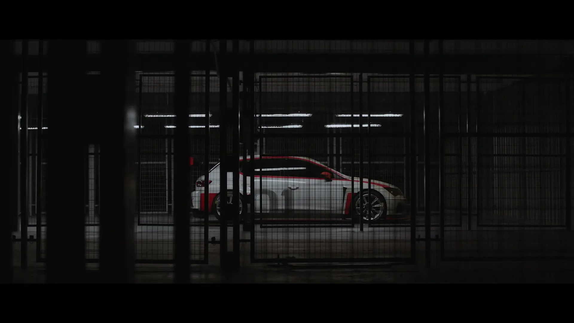 A white race car with red stripes is seen through a metal fence in a dark, industrial garage.