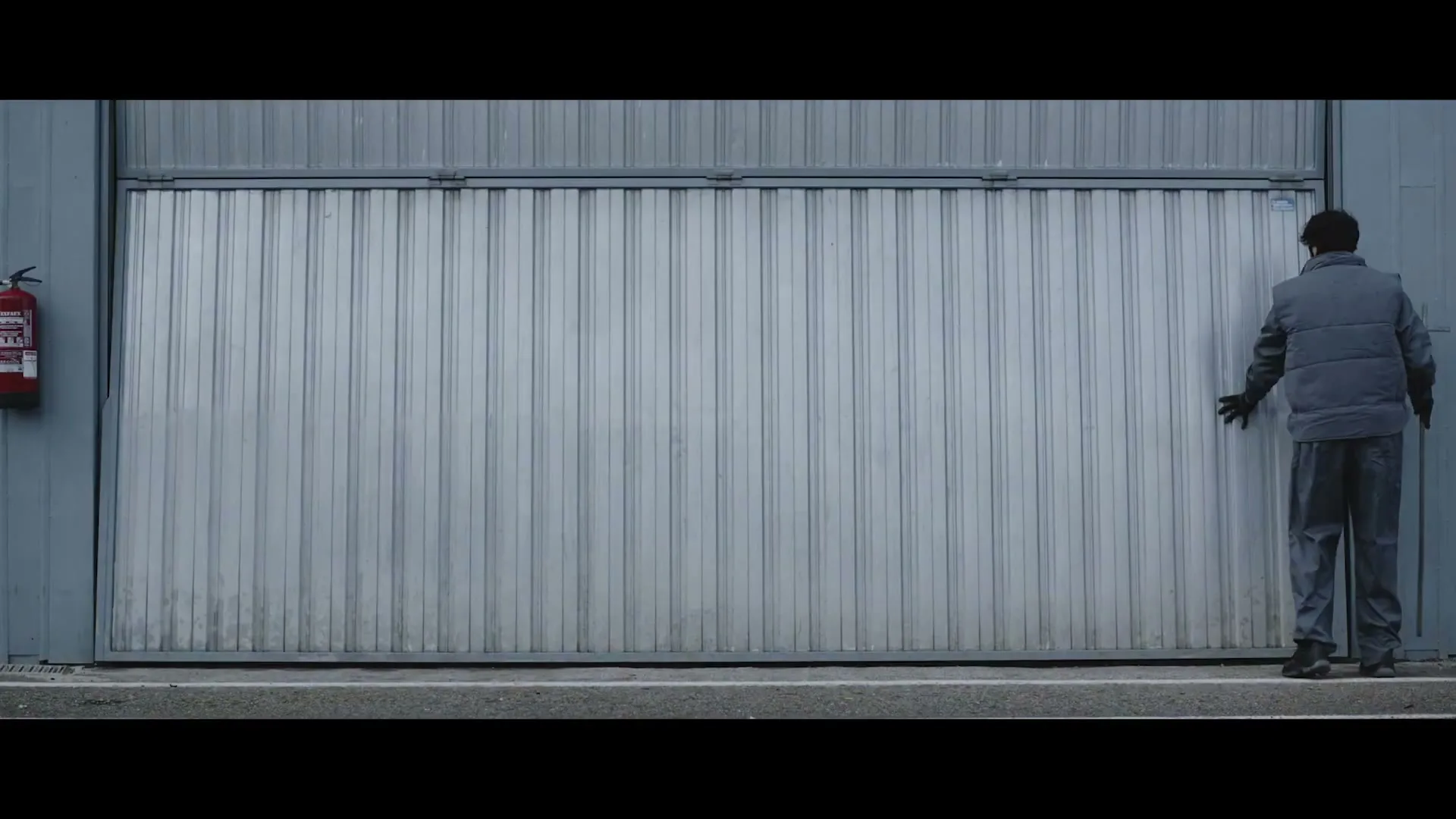 A man in a grey jacket and pants stands next to a large corrugated metal garage door, touching it with his right hand.