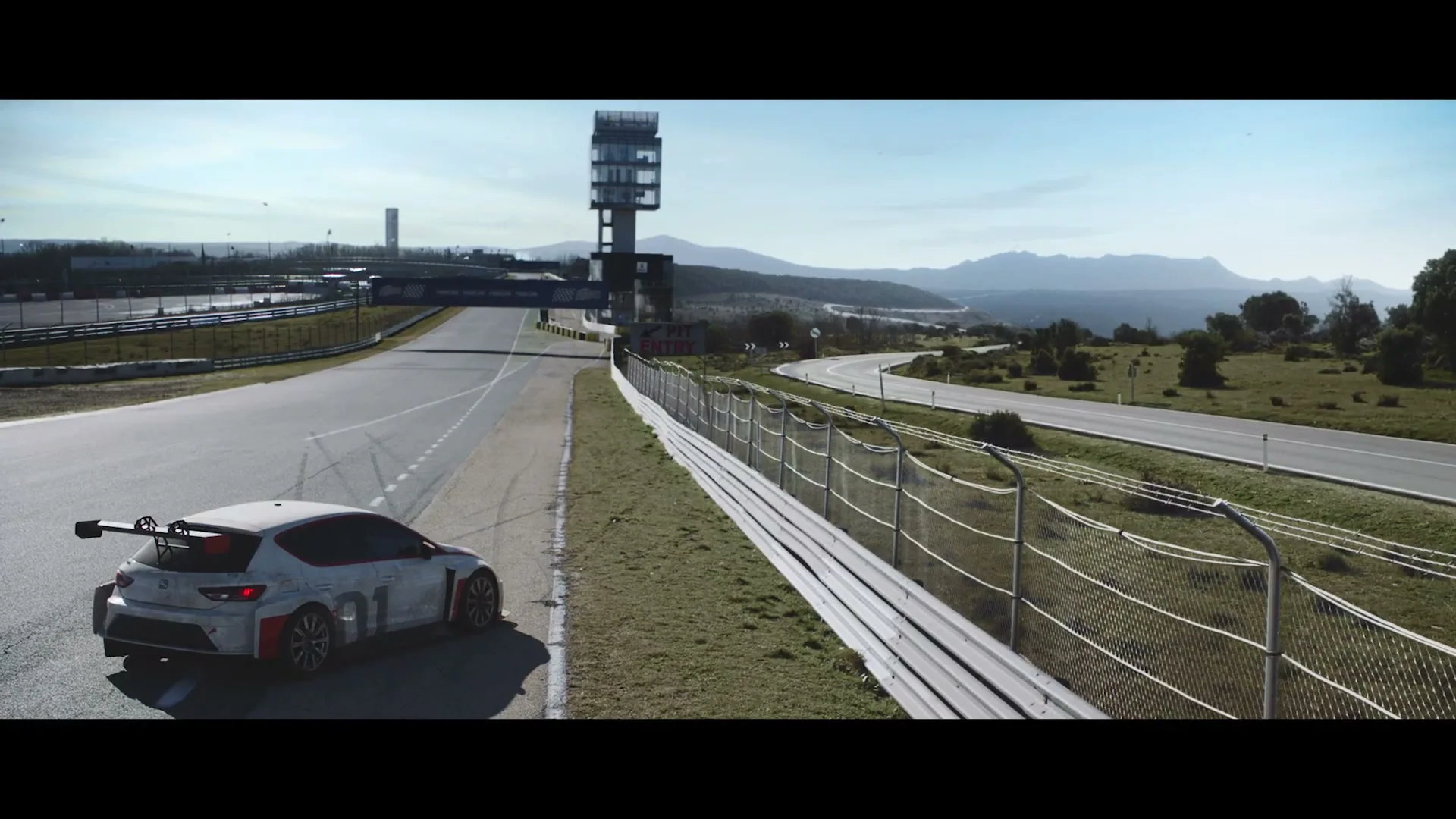 A white race car with red and black accents drives on a race track, with a tall tower and mountains in the background.