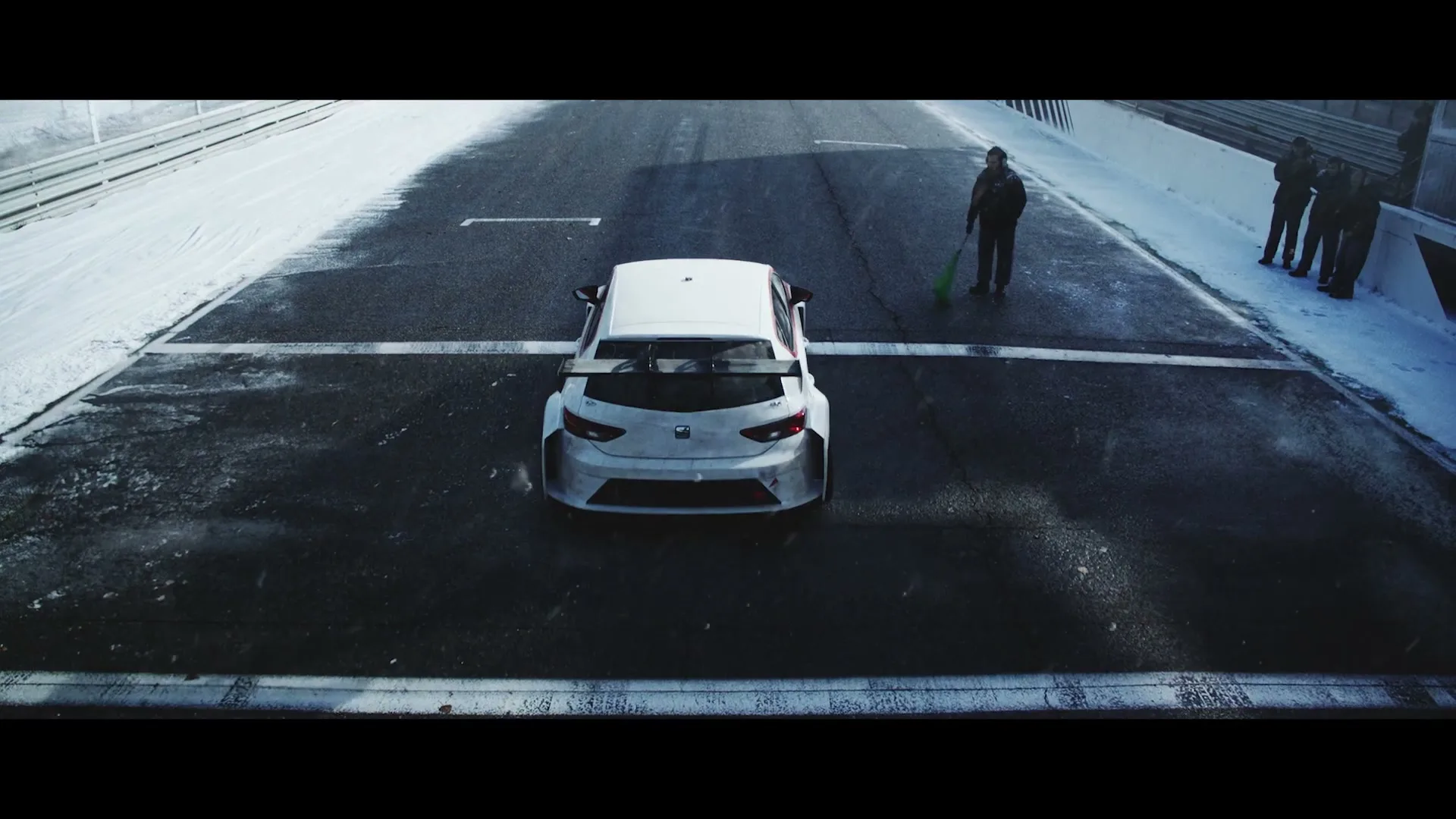 A white race car sits on a snowy track as a man in dark clothing waves a green flag, with other people watching in the background.