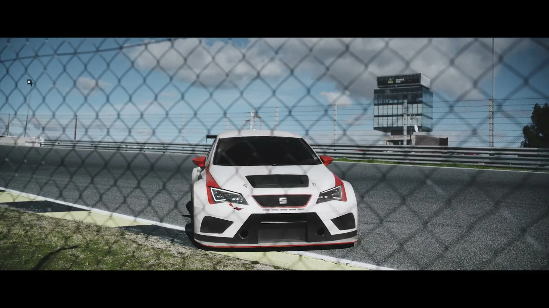 A white race car with red accents is seen through a chain-link fence on a race track under a cloudy sky.