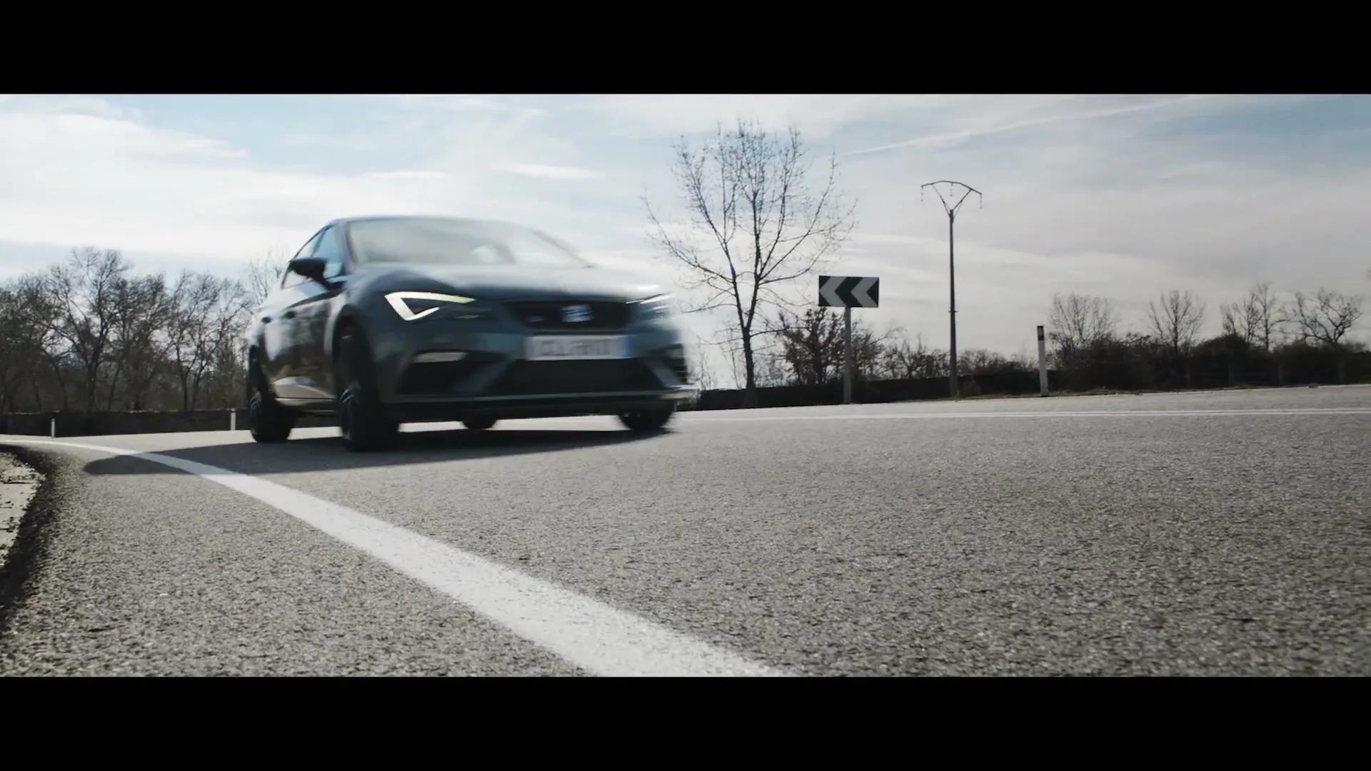 A dark grey Seat car drives quickly around a bend on a rural road under a bright sky.