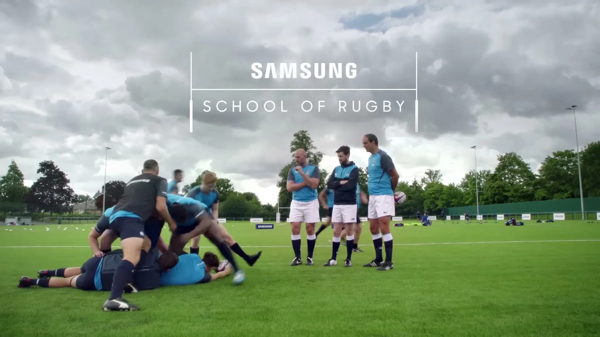 A group of men in blue and black rugby uniforms practice on a green field under a cloudy sky, with some men tackling and others observing.