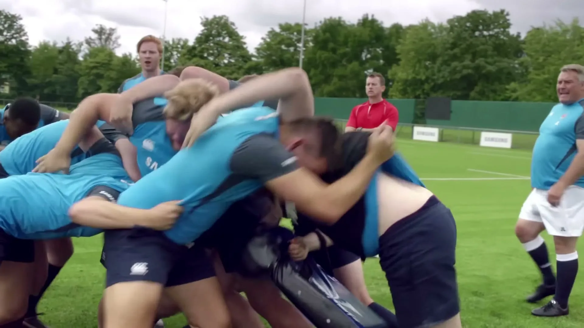 A group of male rugby players in light blue and dark blue uniforms are in a scrum on a green grass field, with coaches watching in the background.
