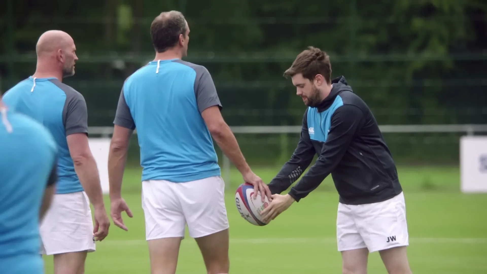 A young man in a black and blue hoodie and white shorts receives a rugby ball from an older man in a blue shirt and white shorts on a green field.