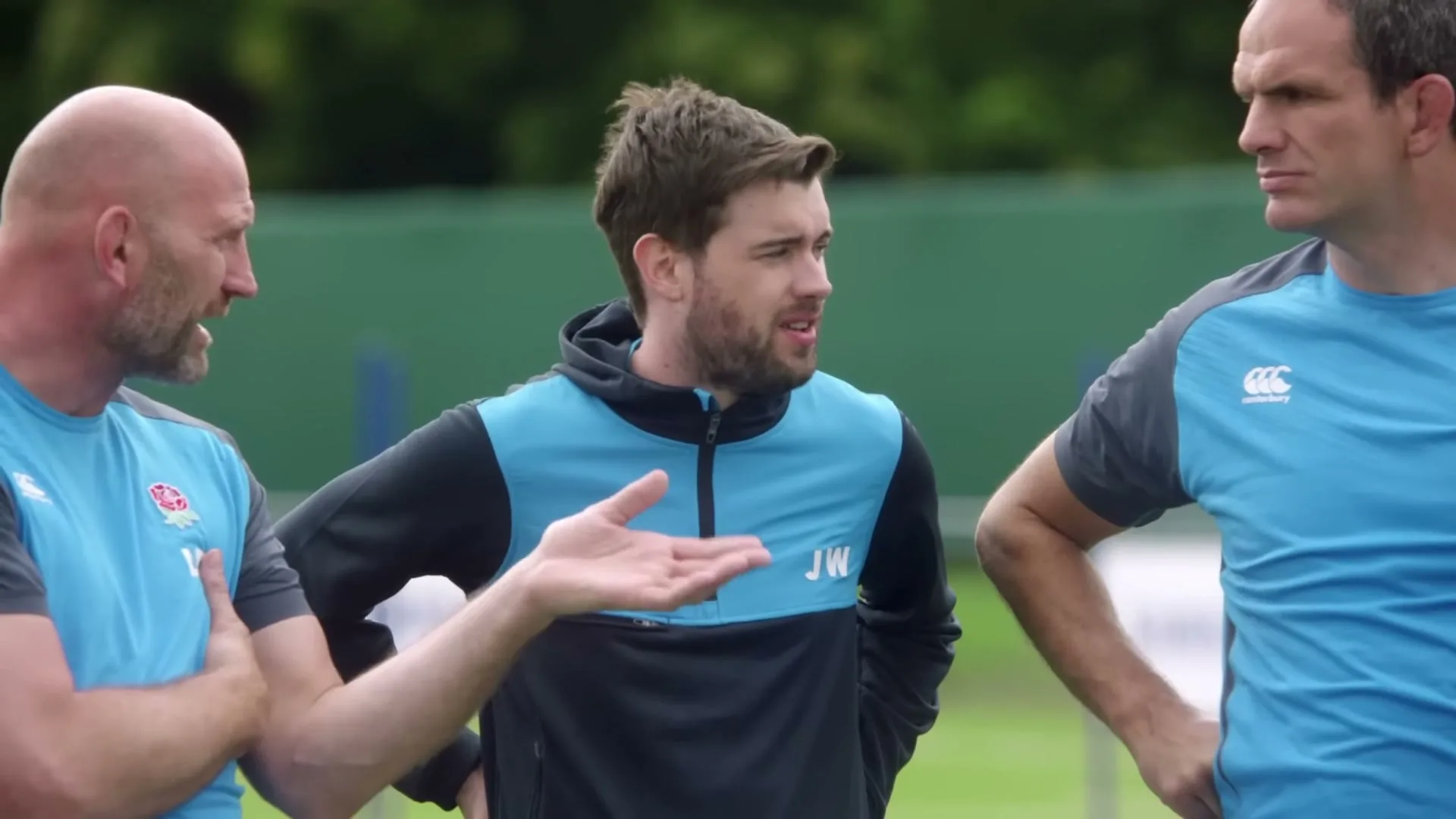Three men in rugby gear stand on a field, with the man on the left gesturing as he speaks to the man in the middle, who listens intently.
