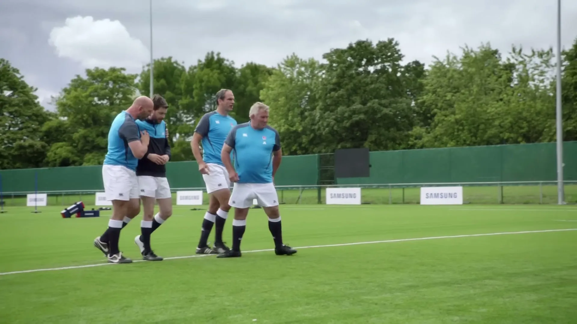 Four male rugby players in blue and white uniforms walk across a green artificial turf field with trees in the background under a cloudy sky.