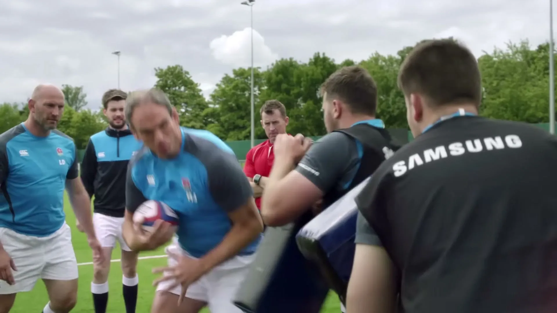 A male rugby player in a blue shirt and white shorts holds a rugby ball and demonstrates a move to other players on a green field.
