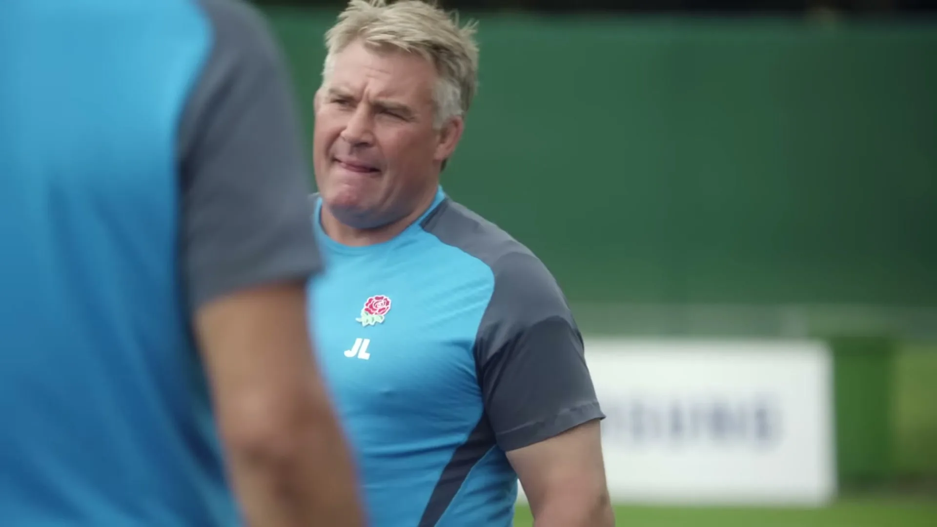 A male rugby coach in a blue and grey shirt looks on with a concerned expression during a practice session on a green field.