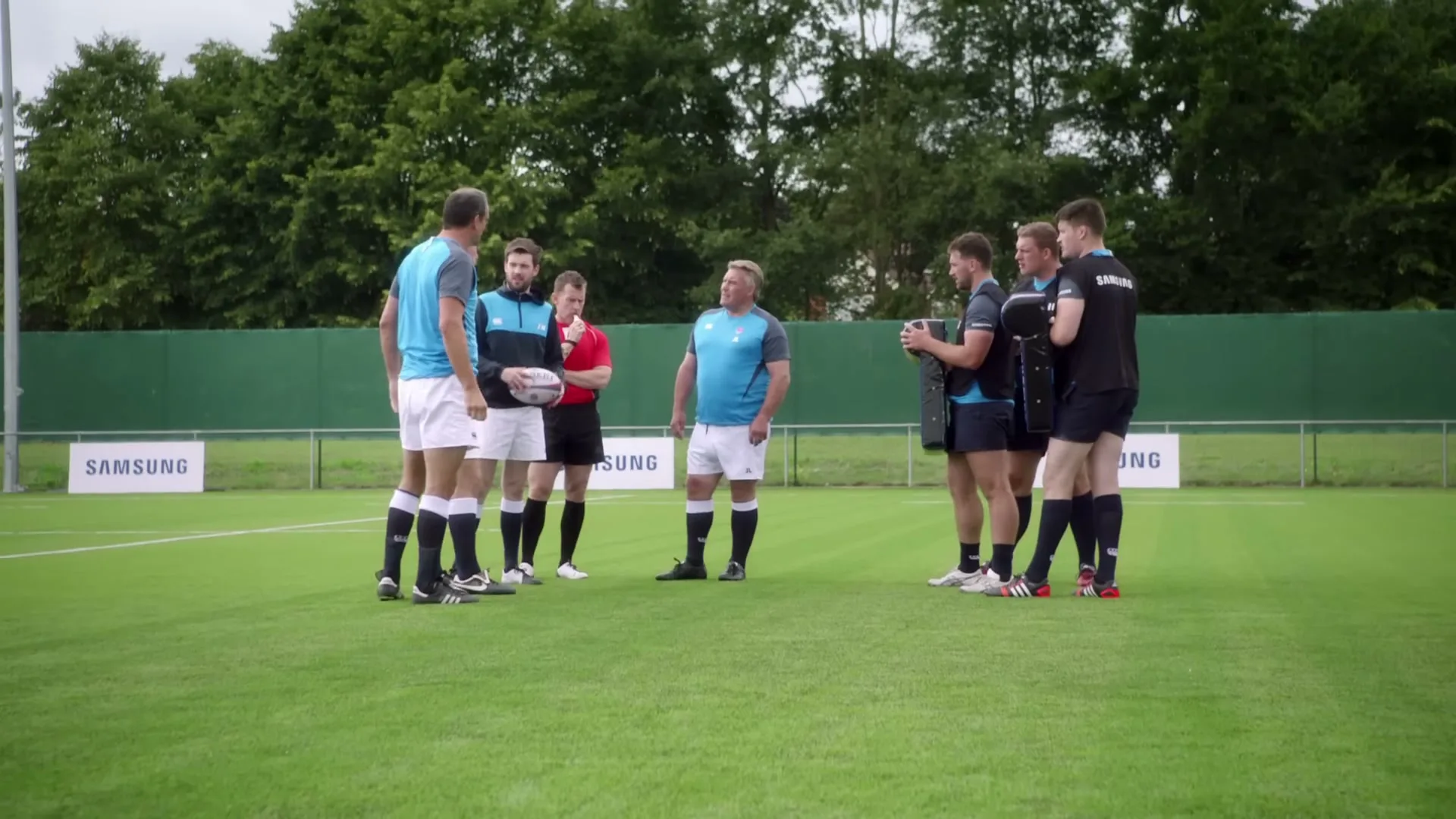 A group of male rugby players and a referee stand on a green field, discussing something during practice.