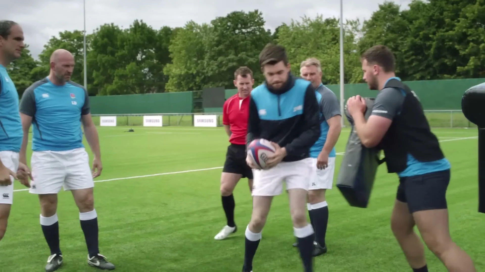 A young man in a blue and black rugby shirt runs with a rugby ball, practicing with a group of men on a green field.