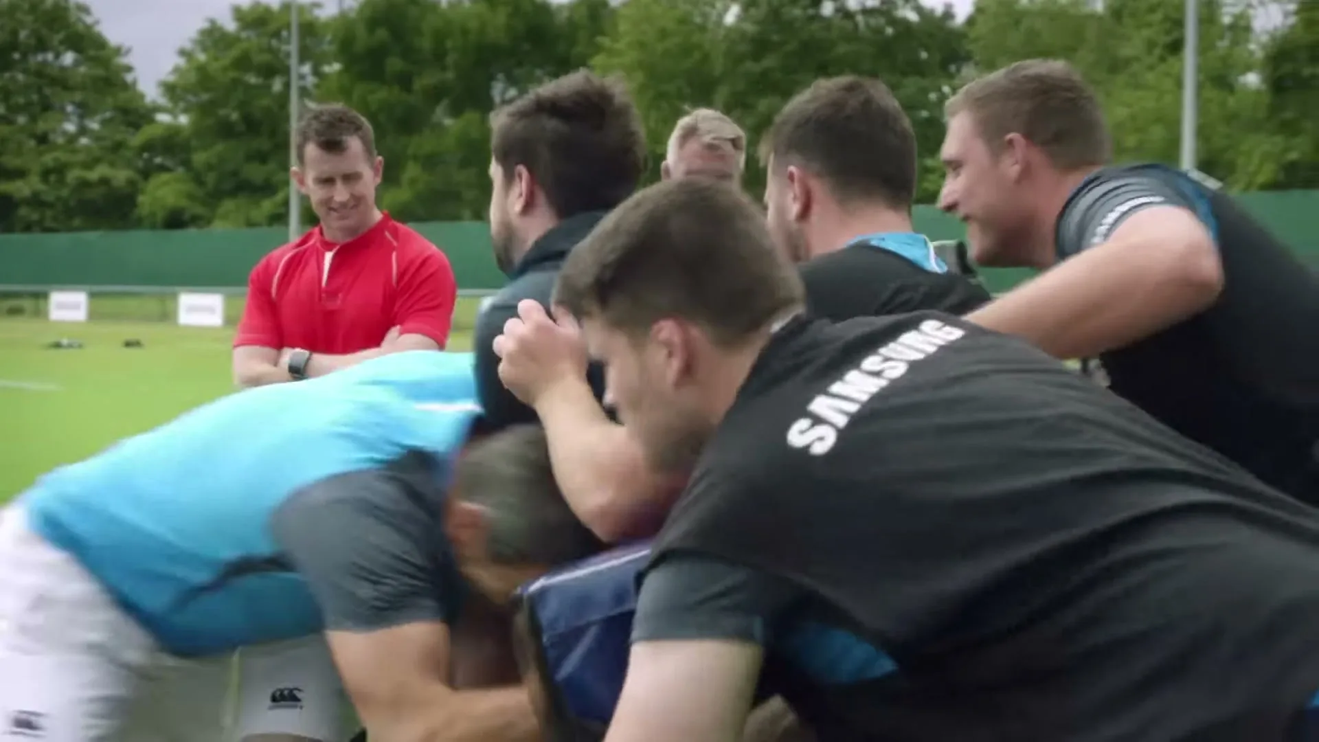 A male rugby coach in a red shirt watches a group of male rugby players in black and blue shirts practicing a scrum on a green field.