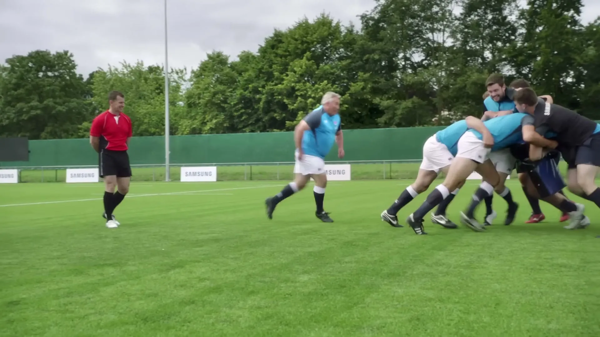 A rugby team in blue and white uniforms practices a scrum on a green field, while a referee in red and black watches them.
