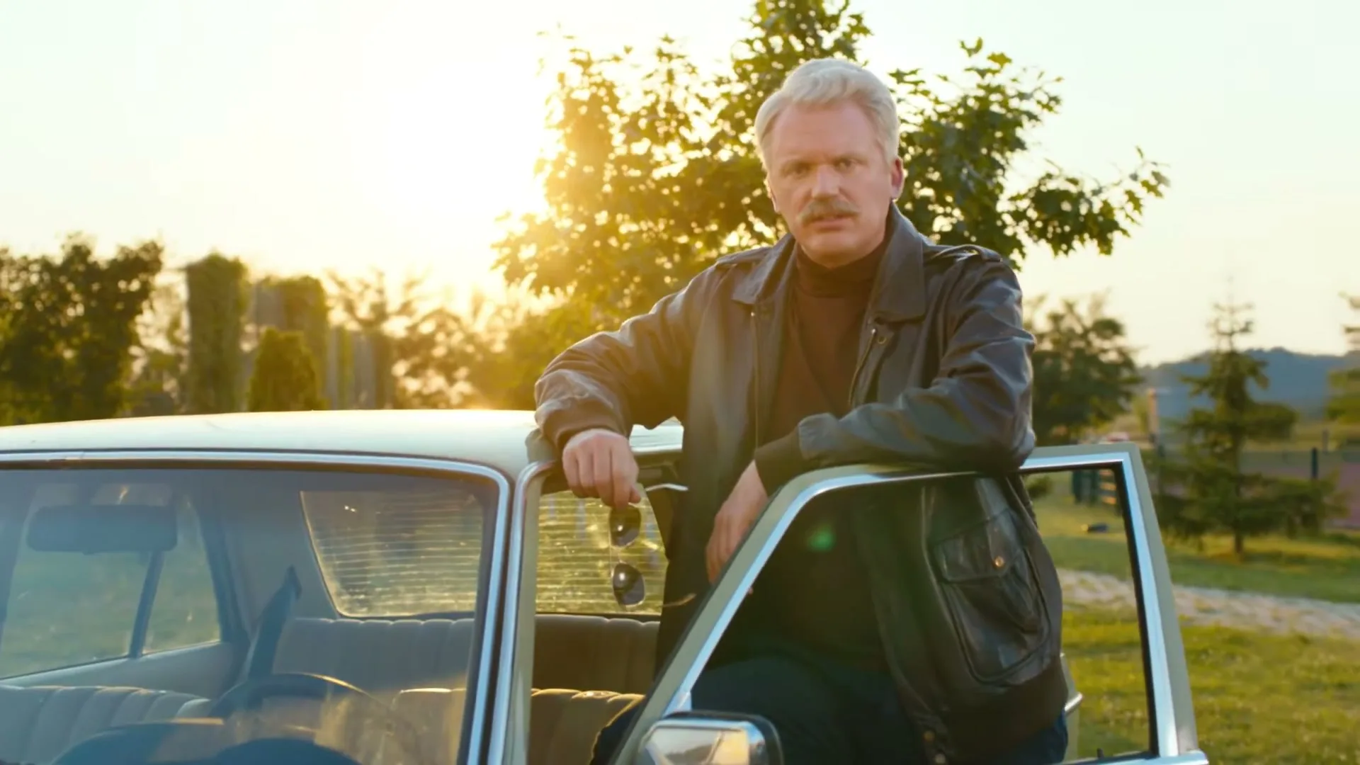 A middle-aged man with a mustache and a leather jacket leans against a vintage car, holding sunglasses, at golden hour in a rural setting.