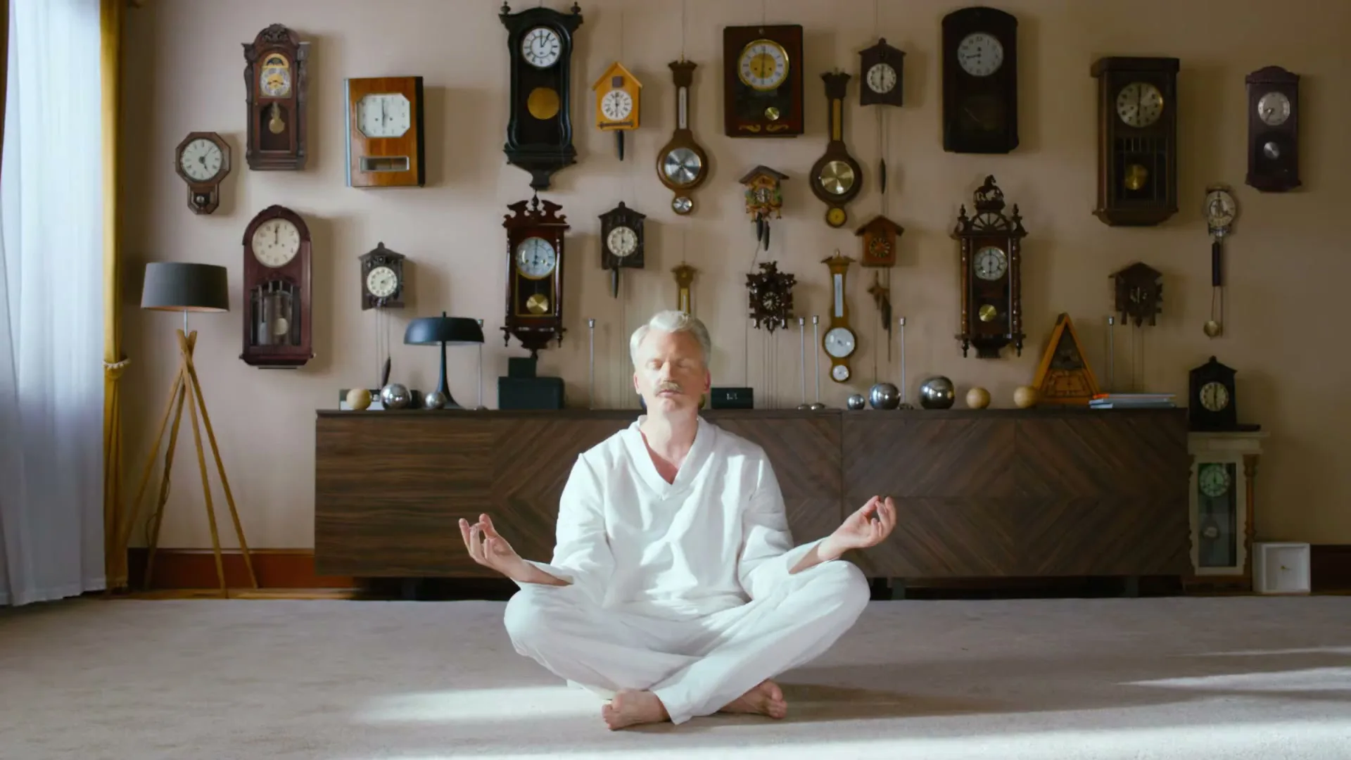 A man with white hair and beard, wearing white clothes, sits cross-legged on the floor, meditating in a room filled with many clocks on the wall.