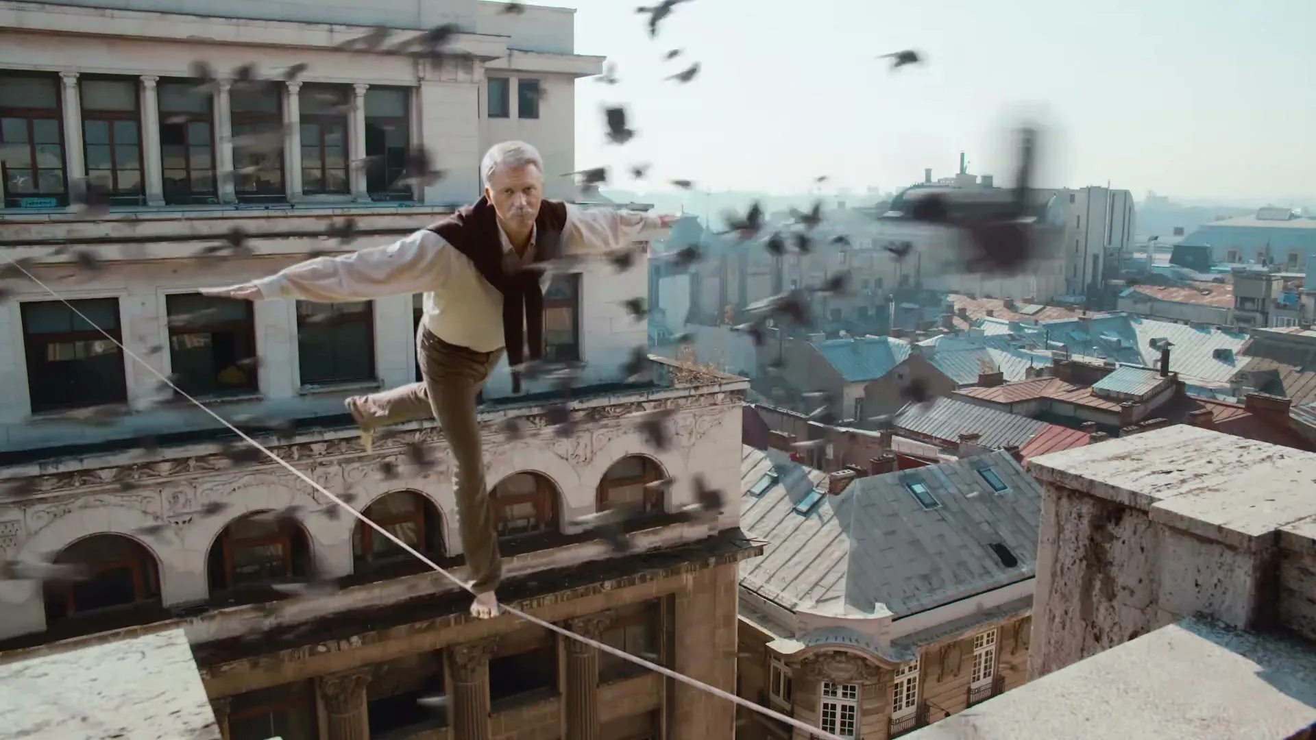 A senior man with a mustache, wearing a white shirt and a sweater, walks barefoot on a tightrope between two buildings, high above a city.
