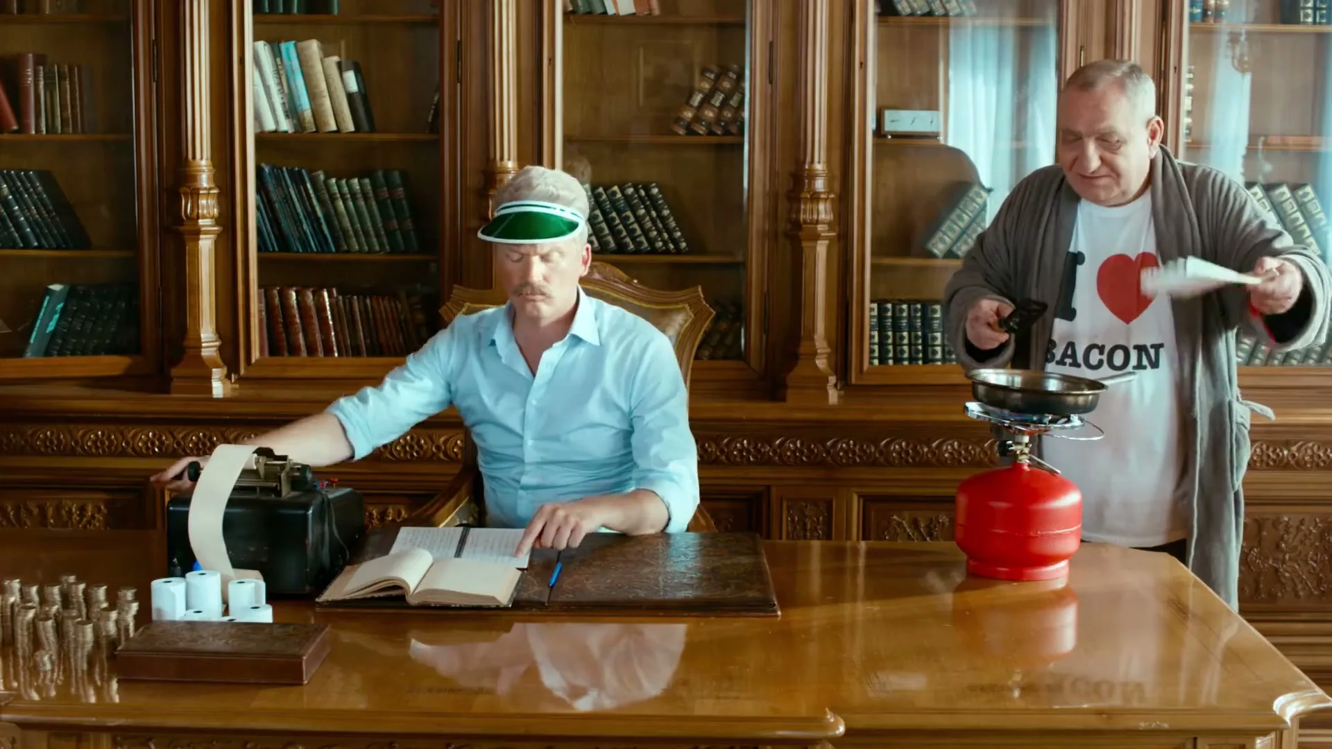 Two men are in a wood-paneled office with bookshelves; one is at a desk with an adding machine, the other stands holding a frying pan with bacon.